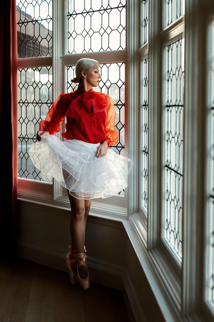 Editorial portrait of a ballerina in a red blouse and tutu by a leaded window
