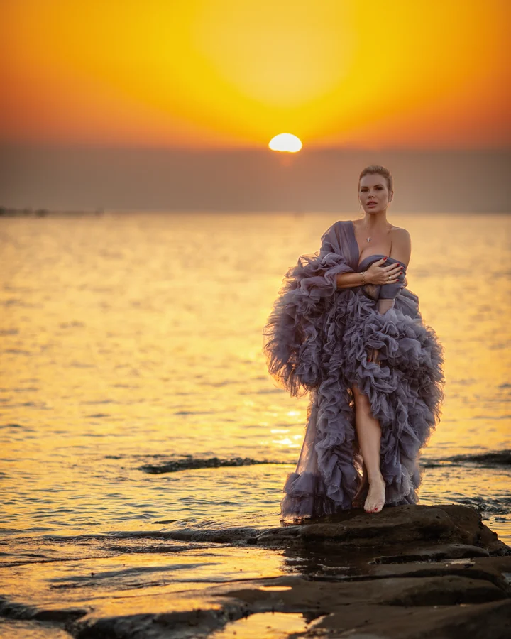 Editorial portrait of a woman in a lilac tulle gown on rocks at sunset over the sea